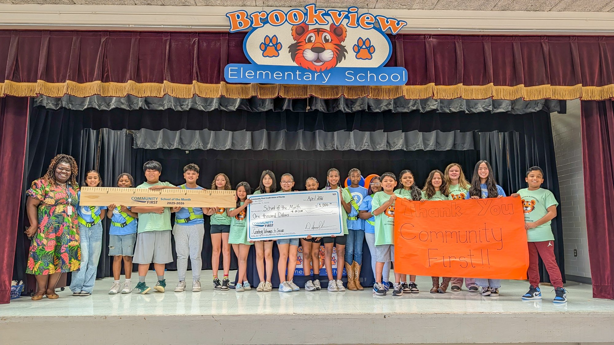 Students on stage with giant ruler, big check and sign.